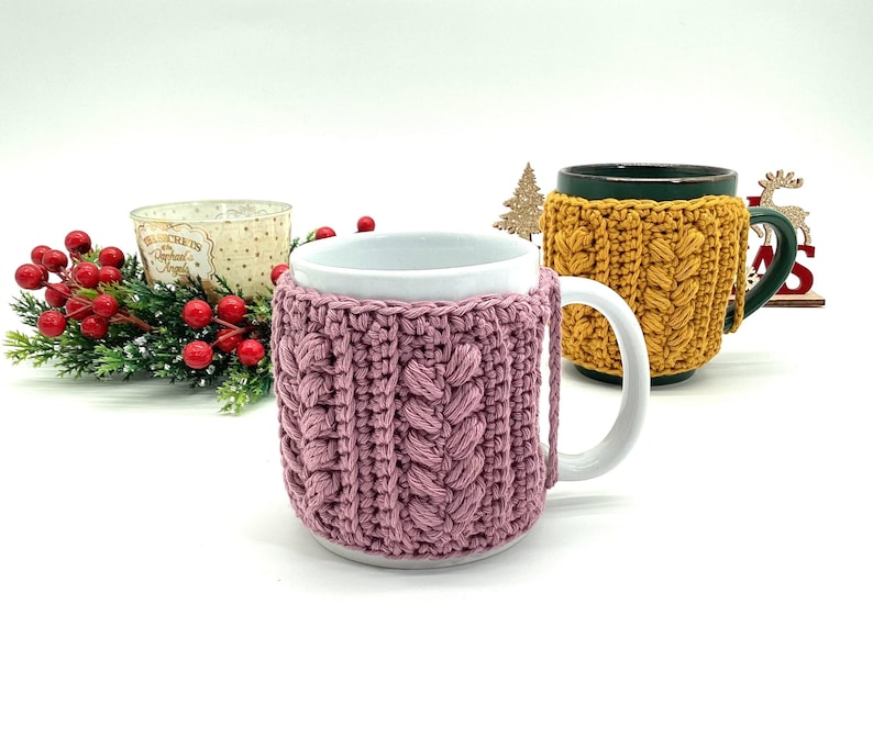May include: Two mugs with crochet cozies. One mug has a pink cozy with a cable knit design, and the other has a yellow cozy. The mugs are on a white surface with holiday decorations in the background.