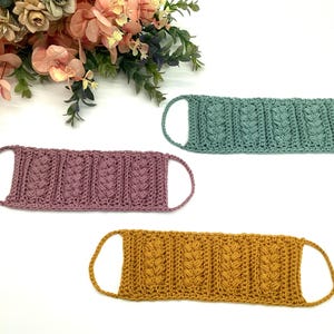 May include: Three handmade crochet face masks in shades of mauve, teal, and mustard yellow. Each mask features a textured, cable-knit design and ear loops. The masks are displayed on a white surface with floral accents in the background.