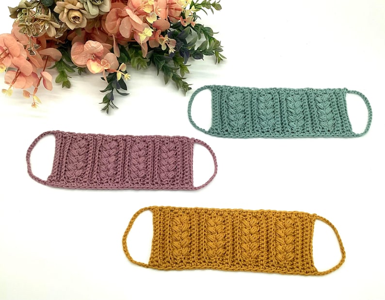 May include: Three handmade crochet face masks in shades of mauve, teal, and mustard yellow. Each mask features a textured, cable-knit design and ear loops. The masks are displayed on a white surface with floral accents in the background.