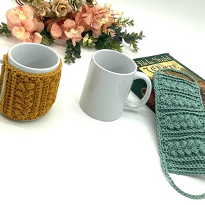 May include: Two white ceramic mugs, one with a mustard yellow knitted cozy. A light teal knitted sleeve is next to a book. Floral arrangement in the background.