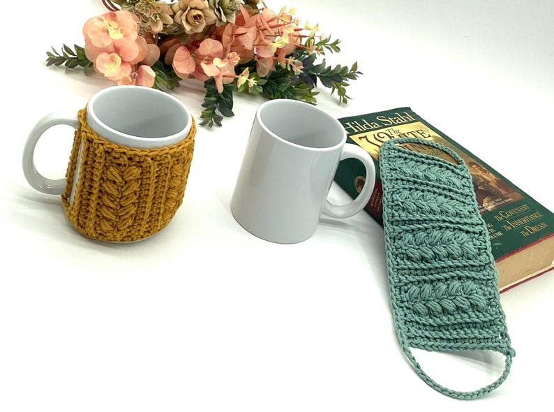 May include: Two white ceramic mugs, one with a mustard yellow knitted cozy. A light teal knitted sleeve is next to a book. Floral arrangement in the background.