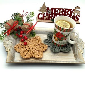 May include: A decorative wooden tray holds a mug with a lemon slice, two crocheted coasters, and Christmas decorations. The mug has a festive design. One coaster is tan, the other is gray. The tray has a distressed finish.