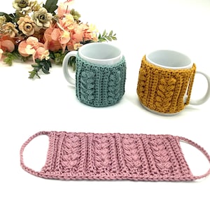 May include: A teal and mustard yellow crochet mug cozy and a pink crochet face mask. The mug cozies have a cable knit design. The face mask has a similar cable knit design with loops for the ears. Artificial flowers are in the background.