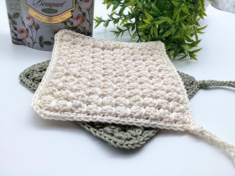 May include: Cream-colored, square crochet bonnet with a textured, raised stitch pattern. A smaller, sage green crochet square is underneath. The bonnet has a long, braided tie. The background includes a tea box and greenery.