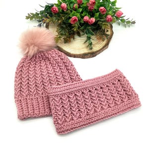 May include: A pink knitted hat with a faux fur pom-pom and a matching neck warmer. Both items feature a textured, woven pattern. The set is presented on a white surface, with a floral arrangement in the background.