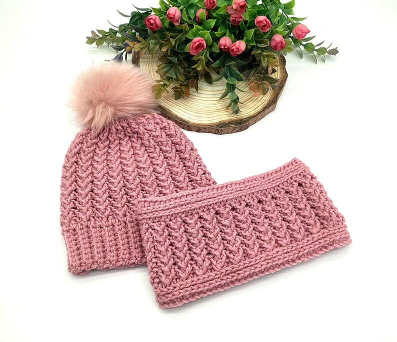 May include: A pink knitted hat with a faux fur pom-pom and a matching neck warmer. Both items feature a textured, woven pattern. The set is presented on a white surface, with a floral arrangement in the background.