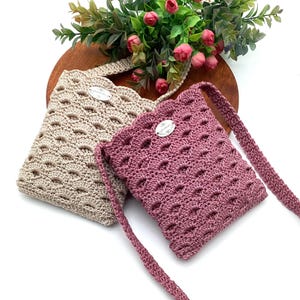May include: Two handmade crochet bags in beige and mauve. The bags have a scalloped edge and a decorative oval tag. The bags have a repeating oval pattern and a strap. The bags are on a wooden surface with flowers.