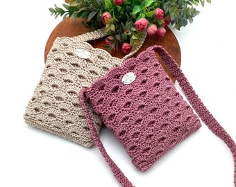 May include: Two handmade crochet bags in beige and mauve. The bags have a scalloped edge and a decorative oval tag. The bags have a repeating oval pattern and a strap. The bags are on a wooden surface with flowers.