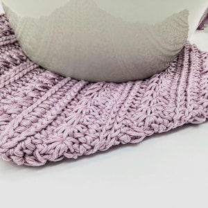 May include: A close-up of a lavender crocheted coaster with a white ceramic bowl on top.
