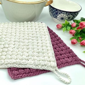 Może przedstawiać: Dwa szydełkowe uchwyty na garnki, jeden biały, a drugi różowy, z fakturowanym, puszystym wzorem. Uchwyty mają pętlę do łatwego zawieszenia.