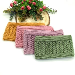May include: Four knitted headbands in shades of gold, pink, and green are arranged in a row. Each headband features a textured, cable-knit design. A small arrangement of pink roses sits in the background.