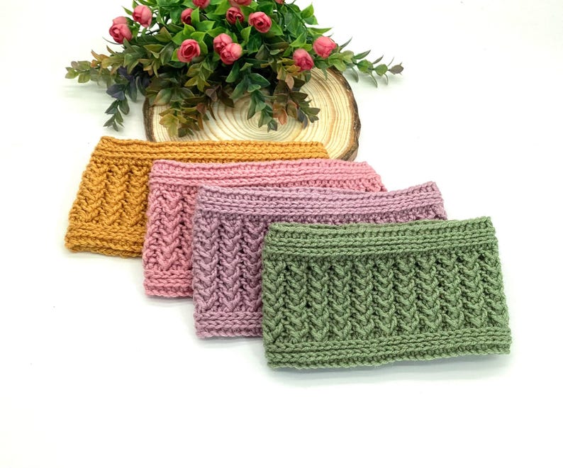 May include: Four knitted headbands in shades of gold, pink, and green are arranged in a row. Each headband features a textured, cable-knit design. A small arrangement of pink roses sits in the background.