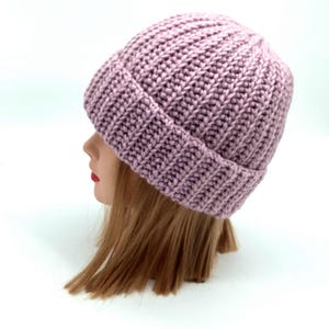 May include: A mauve-colored, ribbed knit beanie hat. The hat is made of a thick yarn and has a folded brim. The hat is shown on a mannequin head with light brown hair and red lipstick.