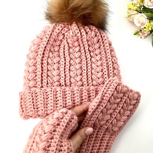 May include: A pink knitted hat with a brown faux fur pom-pom and matching fingerless gloves. The hat and gloves feature a textured, braided design. The set is made of a soft, warm material, ideal for cold weather.