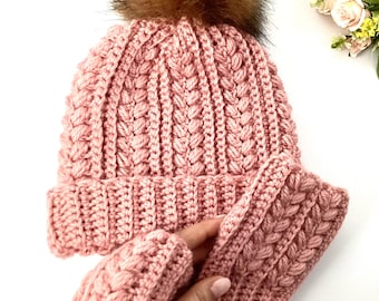 Könnte beinhalten: Ein rosa gestrickter Hut mit einem braunen Kunstfell-Bommel und passenden fingerlosen Handschuhen. Der Hut und die Handschuhe haben ein strukturiertes, geflochtenes Design. Das Set ist aus einem weichen, warmen Material gefertigt.