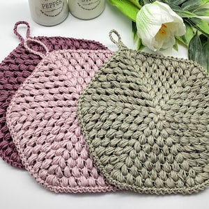 以下が含まれることがあります： ピンク、薄いピンク、緑の3つの編み物鍋つかみ。鍋つかみは丸く、表面がざらざらしています