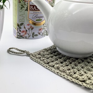 May include: A white ceramic teapot sits on a hand-crocheted, square, light green potholder. The potholder has a loop for hanging. A white ceramic pot with a green plant and a tea tin with floral designs are in the background.