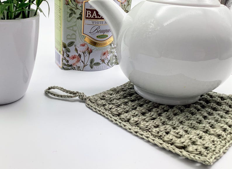 May include: A white ceramic teapot sits on a hand-crocheted, square, light green potholder. The potholder has a loop for hanging. A white ceramic pot with a green plant and a tea tin with floral designs are in the background.
