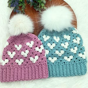 Op de afbeelding: Twee gebreide mutsen met witte pompoenen. De ene muts is roze met witte hartjes en de andere is blauw met witte hartjes.