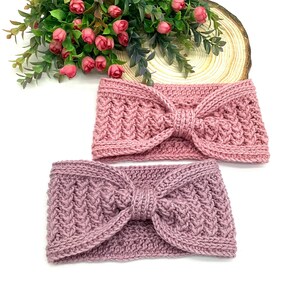May include: Two knitted headbands in shades of pink and mauve. Each headband features a textured pattern and a knotted bow detail at the centre. The headbands are displayed on a white surface with a floral arrangement in the background.