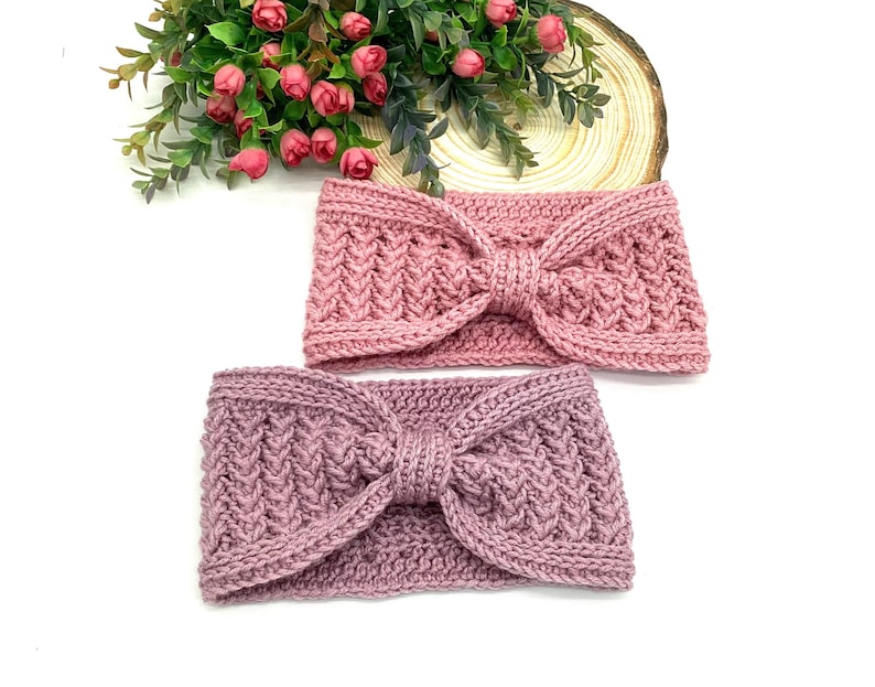 May include: Two knitted headbands in shades of pink and mauve. Each headband features a textured pattern and a knotted bow detail at the centre. The headbands are displayed on a white surface with a floral arrangement in the background.