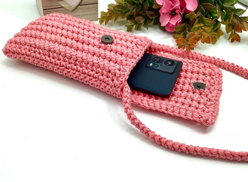May include: A pink crocheted phone case with a black phone inside and a long pink strap.