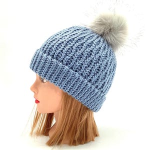 Peut inclure: Un bonnet tricoté bleu clair avec un motif côtelé et un pompon gris duveteux sur le dessus. Le chapeau est présenté sur une tête de mannequin. C'est un accessoire d'hiver.