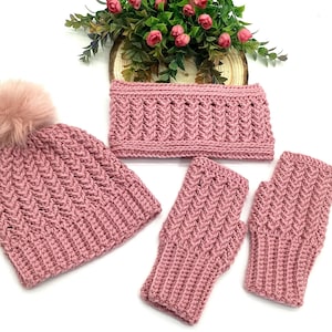 May include: A hand-knitted winter set in a dusty rose hue. The set comprises a beanie with a faux fur pom-pom, a neck warmer, and fingerless gloves. The items are displayed on a white surface, with a floral arrangement in the background.