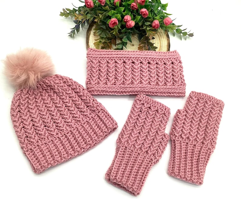 May include: A hand-knitted winter set in a dusty rose hue. The set comprises a beanie with a faux fur pom-pom, a neck warmer, and fingerless gloves. The items are displayed on a white surface, with a floral arrangement in the background.