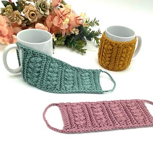 May include: Three handmade crochet mug cozies in teal, mustard yellow, and dusty rose. The cozies feature a textured cable knit design. A white mug is partially covered by the teal cozy. The mustard yellow cozy is wrapped around a white mug. The dusty rose cozy is lying flat.