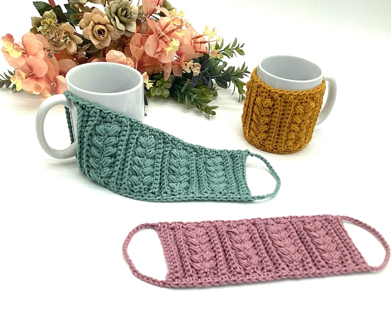 May include: Three handmade crochet mug cozies in teal, mustard yellow, and dusty rose. The cozies feature a textured cable knit design. A white mug is partially covered by the teal cozy. The mustard yellow cozy is wrapped around a white mug. The dusty rose cozy is lying flat.