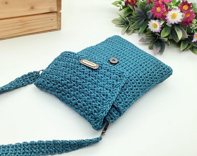 Pu&ograve; includere: Borsa a tracolla all'uncinetto color teal con chiusura a patta e tracolla lunga. La borsa presenta una piccola etichetta rettangolare con testo. &Egrave; realizzata in un materiale strutturato e intrecciato e ha una chiusura magnetica.