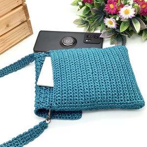 Pu&ograve; includere: Borsa a tracolla all'uncinetto color teal con cinturino abbinato. La borsa ha una forma quadrata e una tasca che contiene un telefono bianco. Uno smartphone nero poggia sulla parte superiore della borsa. La borsa &egrave; realizzata in materiale a maglia strutturato.