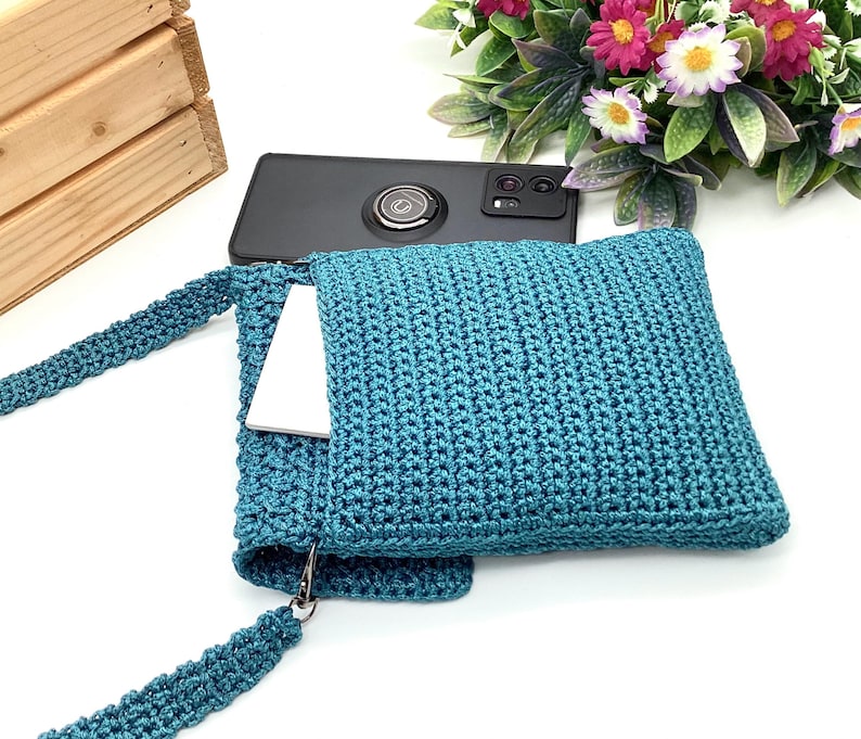 Pu&ograve; includere: Borsa a tracolla all'uncinetto color teal con cinturino abbinato. La borsa ha una forma quadrata e una tasca che contiene un telefono bianco. Uno smartphone nero poggia sulla parte superiore della borsa. La borsa &egrave; realizzata in materiale a maglia strutturato.