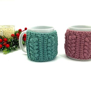 May include: Two white mugs with crocheted cozies. One cozy is teal, the other is mauve. The cozies have a cable knit design. A candle and holiday greenery are in the background.
