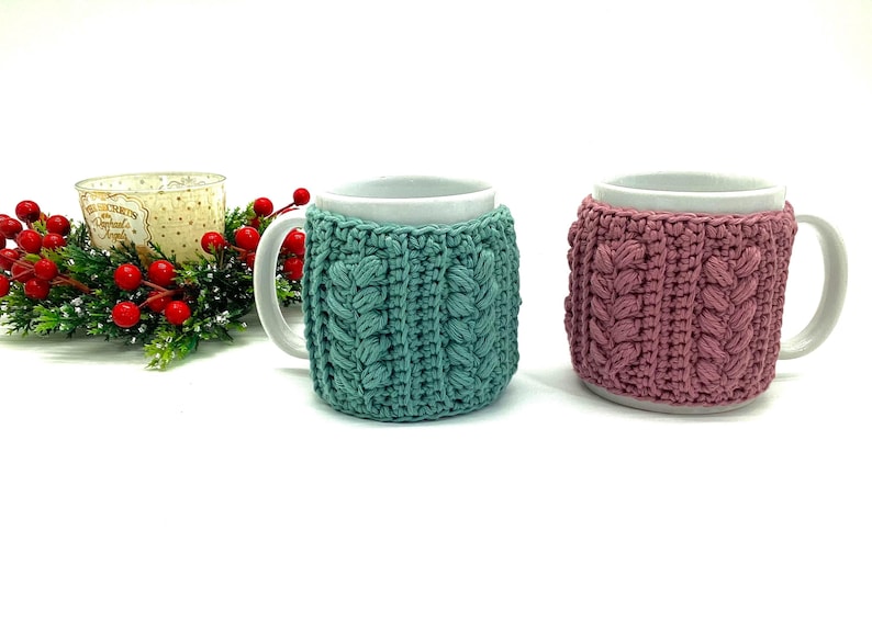 May include: Two white mugs with crocheted cozies. One cozy is teal, the other is mauve. The cozies have a cable knit design. A candle and holiday greenery are in the background.