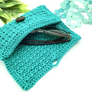 May include: A teal crocheted sunglasses case with a wooden button closure. The case is open and a pair of sunglasses are inside.