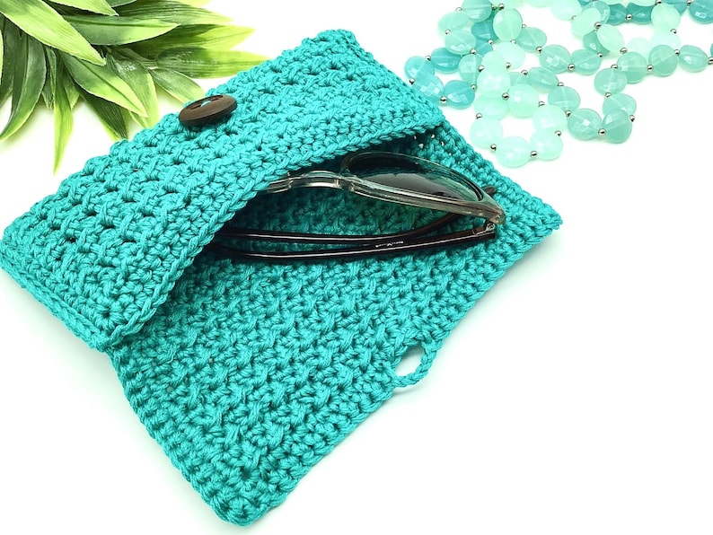 May include: A teal crocheted sunglasses case with a wooden button closure. The case is open and a pair of sunglasses are inside.