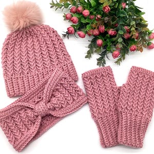 May include: A knitted set in a dusty rose hue, comprising a beanie with a faux fur pom-pom, a headband with a bow, and fingerless gloves. The items feature a textured, chevron-style pattern. The set is designed for warmth and style.