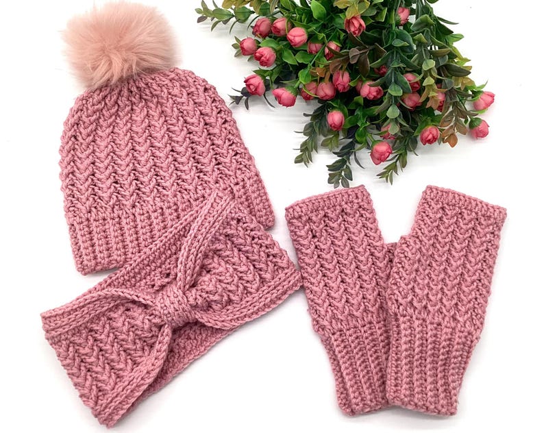 May include: A knitted set in a dusty rose hue, comprising a beanie with a faux fur pom-pom, a headband with a bow, and fingerless gloves. The items feature a textured, chevron-style pattern. The set is designed for warmth and style.