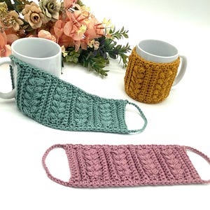 May include: Three handmade crochet mug cozies in teal, mustard yellow, and dusty rose. The cozies feature a textured, raised stitch pattern and are designed to fit around a standard mug. The teal cozy is wrapped around a white mug, and the mustard yellow cozy is on a white mug.