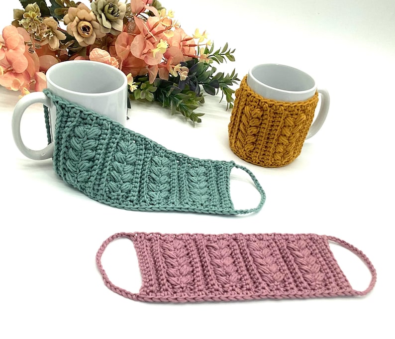 May include: Three handmade crochet mug cozies in teal, mustard yellow, and dusty rose. The cozies feature a textured, raised stitch pattern and are designed to fit around a standard mug. The teal cozy is wrapped around a white mug, and the mustard yellow cozy is on a white mug.