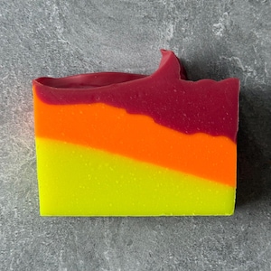 May include: A rectangular bar of handmade soap with a layered design. The top layer is a deep red, followed by orange, and then a bright yellow. The soap has a slightly uneven top edge, suggesting a natural, handcrafted quality.