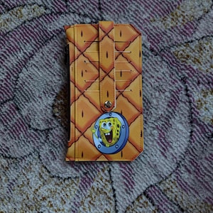 Puede incluir: Un tarjetero amarillo con un patrón repetido de diamantes. Tiene un cierre de solapa con un botón a presión. Un emblema circular de Bob Esponja está en la parte delantera.