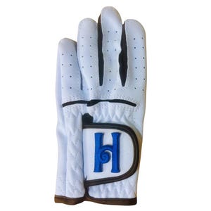 May include: White golf glove with a blue "H" logo on the wrist strap. The glove has black accents and a textured design.