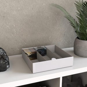 May include: A light grey, three-compartment organiser with a textured exterior. The organiser holds two decks of cards with decorative designs. A small, white, circular object is on the right side. A potted plant is in the background.
