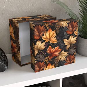 Könnte beinhalten: Eine rechteckige Box mit Deckel, verziert mit einem sich wiederholenden Muster aus Herbstblättern in Orange-, Gelb- und Brauntönen auf dunklem Hintergrund. Die Box ist geöffnet und zeigt ein weißes Innere. Die Box steht auf einer weißen Oberfläche.