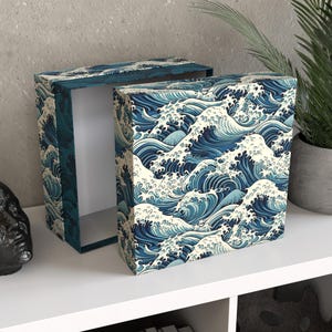 Peut inclure: Deux boîtes décoratives ouvertes avec un motif répété de vagues océaniques bleues et blanches. Les boîtes sont rectangulaires et ont un couvercle à charnière. Les boîtes sont sur une étagère blanche.