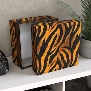 Könnte beinhalten: Geöffnete rechteckige Box mit Deckel, verziert mit einem orange-schwarzen Tigerstreifenmuster. Das Innere der Box ist weiß. Die Box steht auf einer weißen Oberfläche, mit einer Pflanze im Hintergrund.