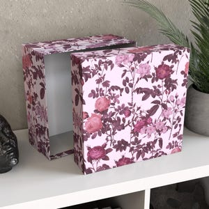 Könnte beinhalten: Zwei rechteckige Aufbewahrungsboxen mit floralem Muster. Die Boxen sind hellrosa mit einem sich wiederholenden Design aus rosa und burgunderfarbenen Rosen, Blättern und Stielen. Eine Box ist geöffnet und zeigt einen weißen Innenraum.
